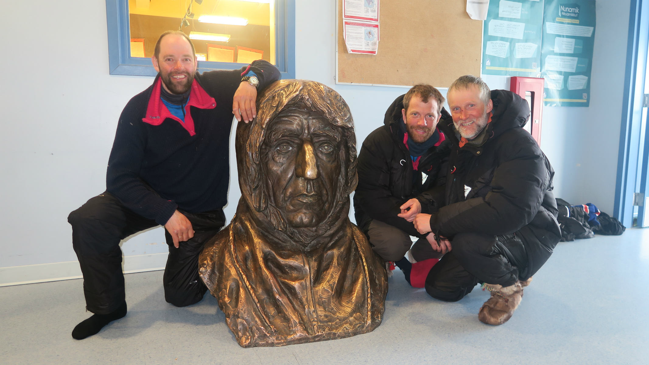Tre menn sitter på huk rundt en byste av Roald Amundsen